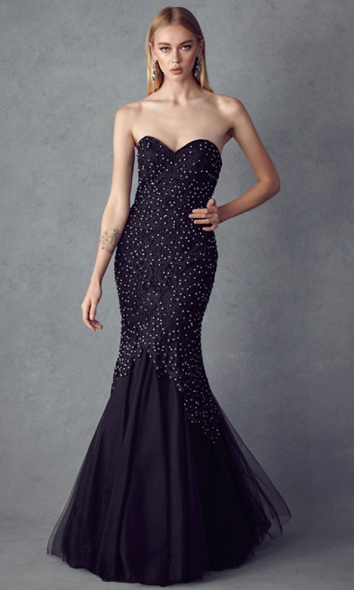 A woman poses against a gray backdrop in the Juliet 644 Long Mermaid Prom Dress, featuring a strapless design, beaded embellishments, and a flared tulle hem—ideal for prom or formal occasions.
